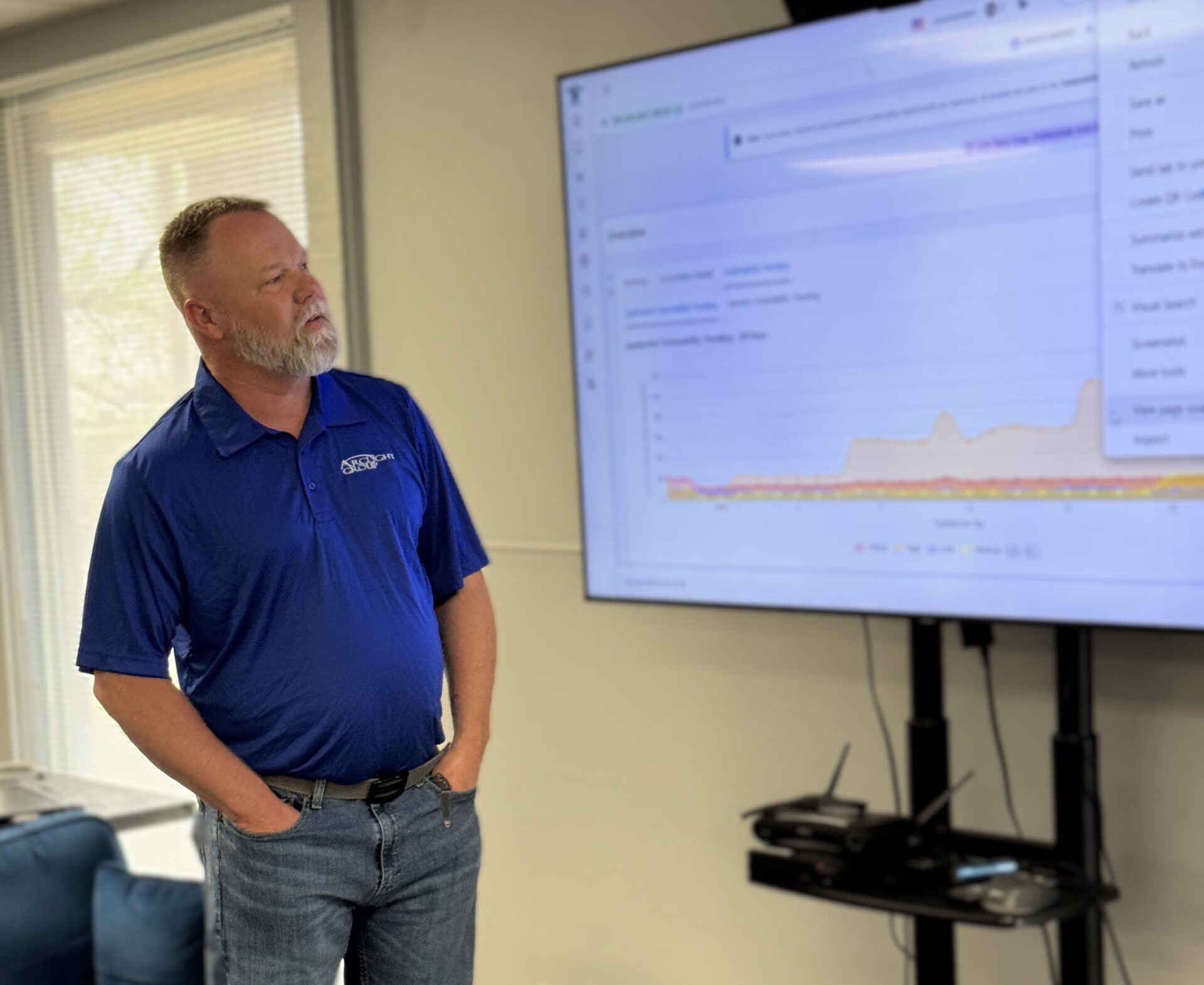 Man in a blue polo presenting beside a large screen with a data chart in an office conference room.