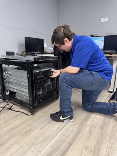 Technician kneeling beside an open server rack, adjusting hardware in a small tech lab.